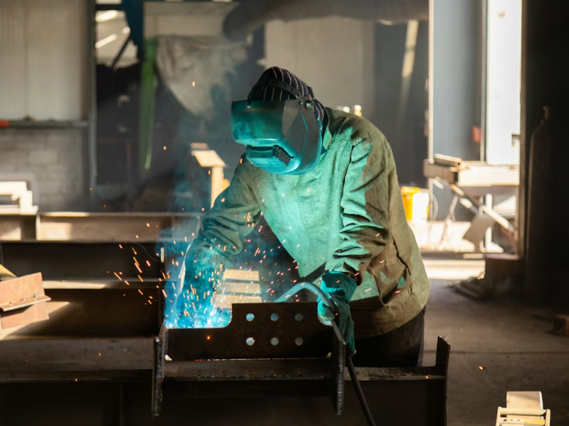 A imagem mostra uma pessoa realizando um processo de soldagem em um ambiente industrial, possivelmente uma fábrica metalmecânica.