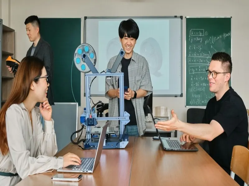 três estudantes conversam entre si sobre parceria entre universidade e indústria e com auxílio de computadores e aparelhos de inovação