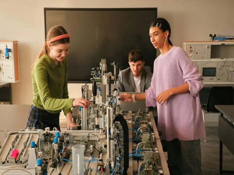 estudantes interagem com projeto robótico em sala de aula, com professor ao fundo avaliando
