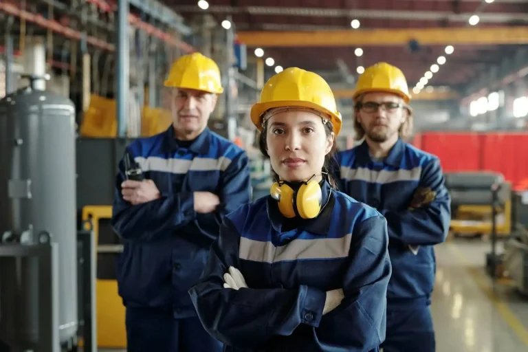 Três trabalhadores em um ambiente industrial, todos usando uniformes azuis e capacetes amarelos, representando trabalho em equipe e segurança no trabalho.