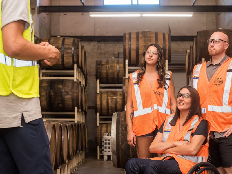 A imagem retrata um grupo diversificado de trabalhadores em uma instalação de produção ou armazenamento, possivelmente uma destilaria, vinícola ou cervejaria, recebendo instruções ou participando de um briefing.