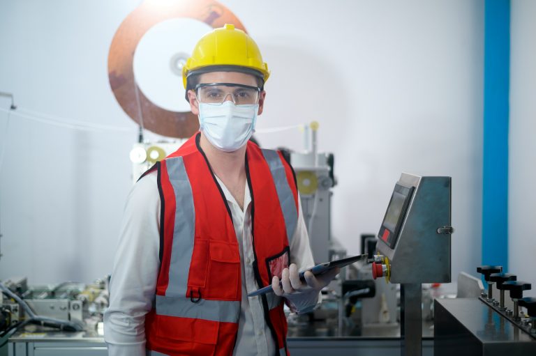 Profissional de segurança no ambiente industrial usando capacete amarelo, máscara, óculos de proteção e colete refletivo, segurando um tablet.