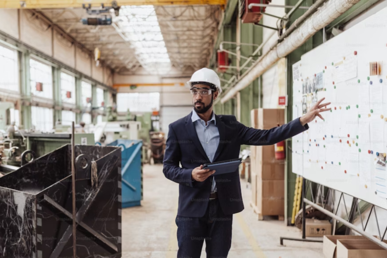 A imagem mostra um profissional da área industrial — usando capacete de segurança, óculos de proteção e traje formal — em uma fábrica ou chão de produção, apontando para um quadro com diagramas e informações organizadas com alfinetes coloridos. Ele segura um tablet, indicando o uso de tecnologia no acompanhamento das operações.