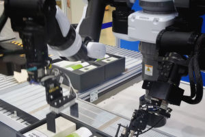 A imagem mostra dois braços robóticos industriais realizando uma operação automatizada em uma linha de montagem. Os robôs, de aparência moderna e com estrutura metálica nas cores preto, branco e cinza, pertencem à marca Kawada, visível em um dos equipamentos.