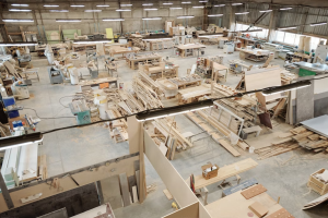 imagem é uma visão ampla e aérea (de cima) do interior de um grande galpão que funciona como uma fábrica ou oficina de marcenaria/móveis, o que é consistente com o nome do arquivo ("automação robótica para PMEs moveleiras.jpg").