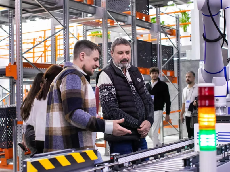 
A imagem retrata um grupo de pessoas em um ambiente que parece ser um armazém automatizado, centro de distribuição ou, mais provavelmente, uma feira/exposição de tecnologia e automação logística e industrial.

Descrição Detalhada:

Pessoas e Interação: O foco está em dois homens em primeiro plano, que parecem ser o especialista (ou vendedor) e um cliente/visitante:

Homem à Esquerda (Mais Jovem): Veste uma camisa xadrez em tons de marrom, azul e branco. Ele está gesticulando com a mão aberta em direção a um equipamento à frente, parecendo explicar ou apresentar a tecnologia ao outro homem.

Homem ao Centro (Mais Velho): Veste uma camisa com detalhes em tricô (possivelmente com um padrão de inverno ou Fair Isle) por baixo de um colete acolchoado preto. Ele ouve atentamente, com uma expressão concentrada.

Grupo de Fundo: Outras pessoas (visitantes ou funcionários) são visíveis ao fundo, observando ou caminhando pela área de exposição.

Tecnologia e Automação: Vários elementos de automação industrial são visíveis:

Linha de Transporte/Esteira: Em primeiro plano, há uma seção de esteira transportadora com rolos metálicos, ladeada por sinalização de segurança em listras amarelas e pretas.

Torre de Sinalização (Andon): À direita, há uma torre de sinalização industrial (ou coluna de luz/Andon), com segmentos luminosos nas cores verde, amarelo e vermelho. Estas luzes são usadas em fábricas e armazéns para indicar o status da máquina ou do processo (verde: operando, amarelo: aviso/parado, vermelho: falha/emergência).

Braço Robótico/Equipamento: No canto superior direito, um pedaço de um braço ou estrutura robótica de cor clara (branca/cinza) é parcialmente visível, ligado à linha de montagem ou transporte.

Ambiente: O ambiente é composto por:

Estruturas de Estocagem: Grandes estruturas de prateleiras metálicas (racks) de cor laranja e cinza são visíveis no fundo, típicas de armazéns de grande altura.

Organização: O local parece muito limpo e organizado, com plantas ornamentais e iluminação clara, reforçando o cenário de uma exposição ou um armazém de última geração (Smart Warehouse).

Interpretação:

A imagem ilustra a Automação de Logística e Processos (Indústria 4.0), onde a tecnologia (robótica, esteiras, sensores e sistemas Andon) está sendo integrada para otimizar o fluxo de trabalho e o gerenciamento de estoque, um tema relevante para a eficiência operacional e a gestão de riscos ESG (especialmente o pilar Ambiental e Social, devido à otimização e segurança).