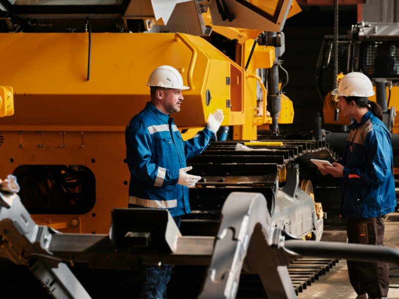 A imagem mostra dois profissionais em um ambiente industrial, possivelmente uma fábrica de máquinas pesadas. Ambos estão usando equipamentos de proteção individual, como capacete branco, óculos de segurança e uniformes industriais.