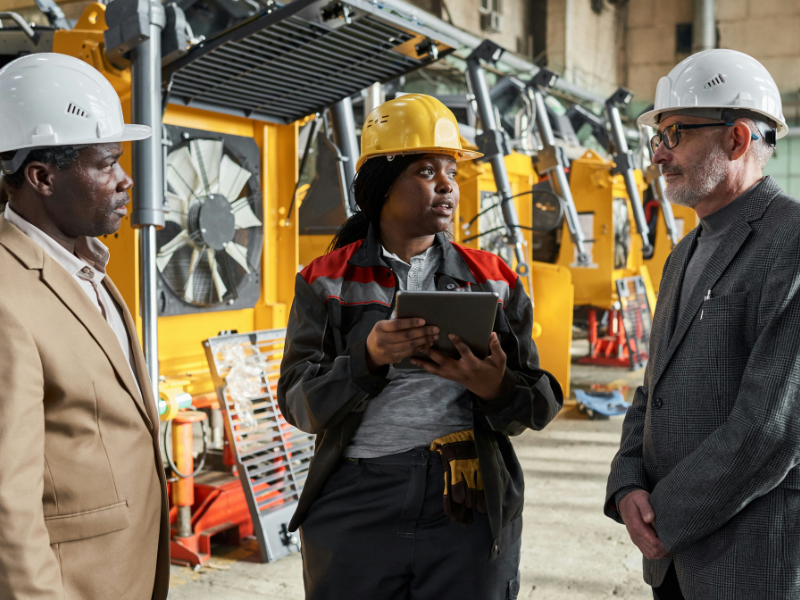 A imagem mostra três pessoas em um ambiente industrial, possivelmente uma fábrica ou canteiro de obras, em frente a maquinário grande e amarelo.