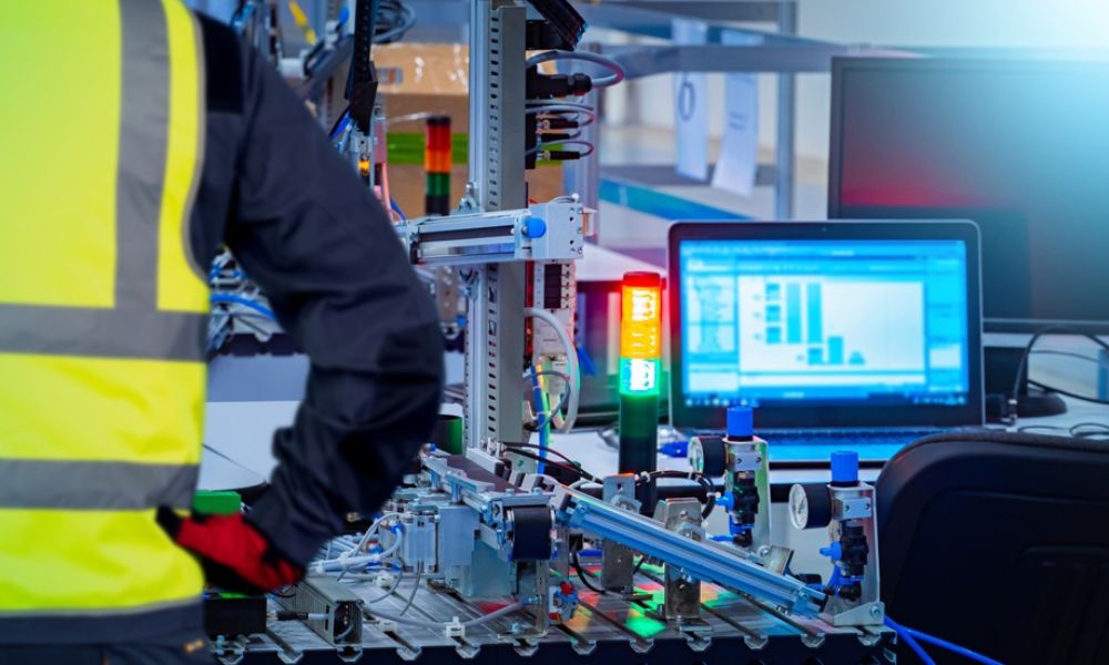 Operador ou técnico vestindo colete de segurança amarelo reflexivo monitorando um sistema industrial automatizado em uma bancada de trabalho.