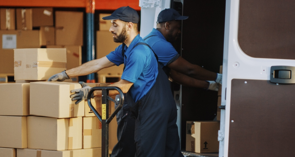 A imagem retrata dois trabalhadores de logística ou entregadores em meio ao processo de carregamento ou descarregamento de mercadorias em um caminhão ou van.