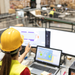 A imagem ilustra uma operária de logística ou engenheira supervisionando a operação de um grande centro de distribuição através de sistemas de computador e monitoramento.