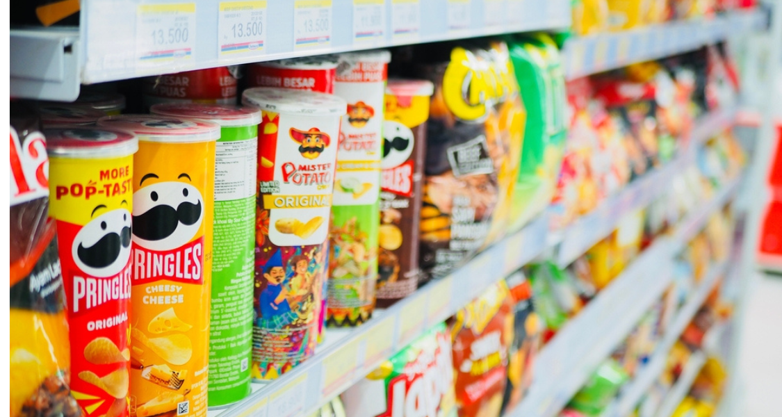 A imagem mostra uma prateleira de supermercado com diversas embalagens de salgadinhos e batatas fritas, em sua maioria do tipo chips em tubo cilíndrico, como as famosas Pringles e Mister Potato. A imagem foca nas cores e nas formas dos produtos, destacando como o design visual influencia o apetite.