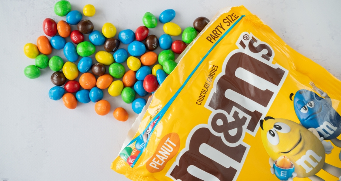 A imagem mostra um pacote de M&M’s amarelo, versão Peanut (amendoim), do tipo Party Size (tamanho festa), parcialmente aberto com várias unidades dos confeitos coloridos espalhadas sobre uma superfície clara.
