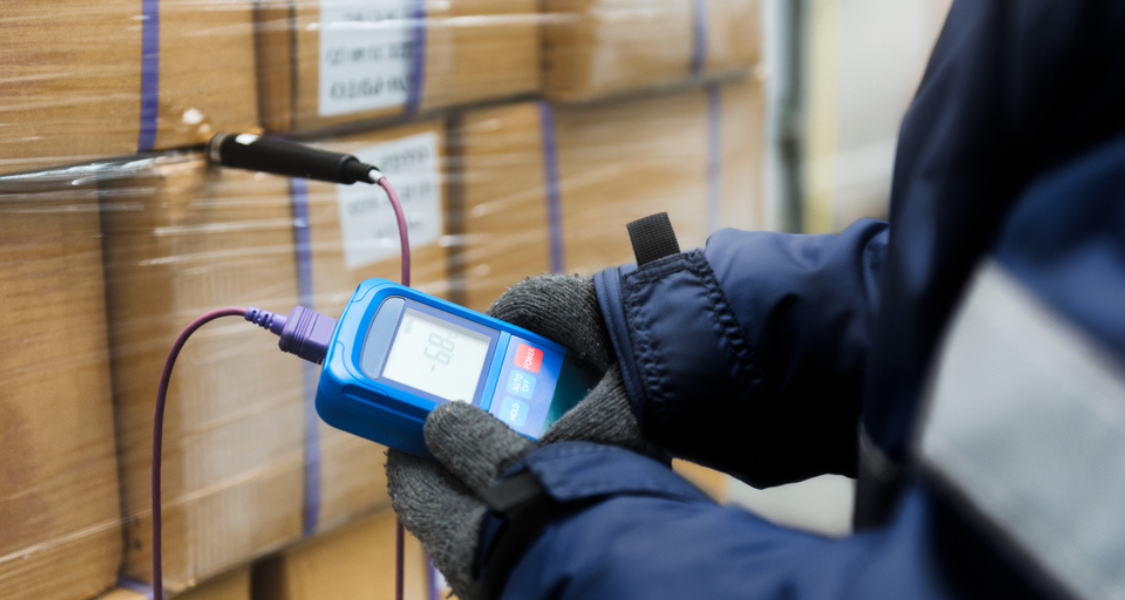 A imagem mostra uma pessoa usando luvas térmicas e jaqueta acolchoada, manipulando um termômetro digital portátil conectado por cabos a uma sonda inserida em uma pilha de caixas embaladas com filme plástico.