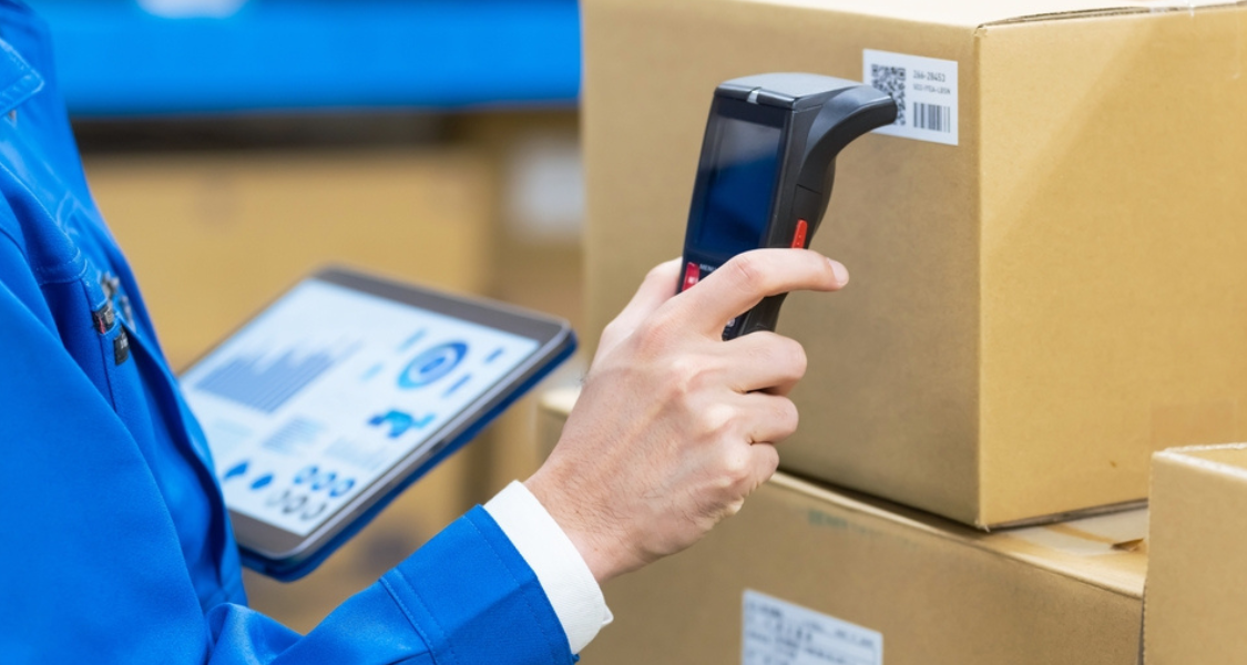 A imagem mostra uma pessoa vestida com uniforme azul, segurando um leitor de código de barras e um tablet enquanto realiza o escaneamento de uma etiqueta em uma caixa de papelão.