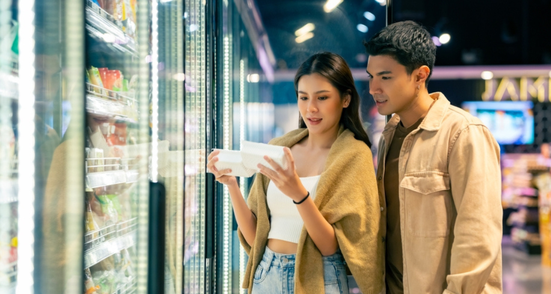 A imagem mostra um casal jovem em um supermercado, em frente a um corredor de freezers com portas de vidro iluminadas.