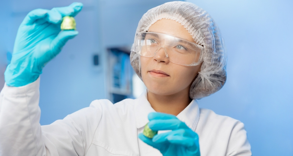 A imagem retrata uma profissional em ambiente de laboratório, vestindo equipamentos de proteção individual. Ela usa jaleco branco, luvas azuis, óculos de proteção e touca descartável, indicando um contexto de trabalho científico ou industrial controlado.