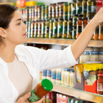 A imagem mostra uma mulher em um supermercado, em frente a uma prateleira repleta de molhos, conservas e produtos em vidro. Ela segura um frasco com tampa verde em uma das mãos enquanto estende o braço para pegar ou comparar outro produto semelhante na prateleira.