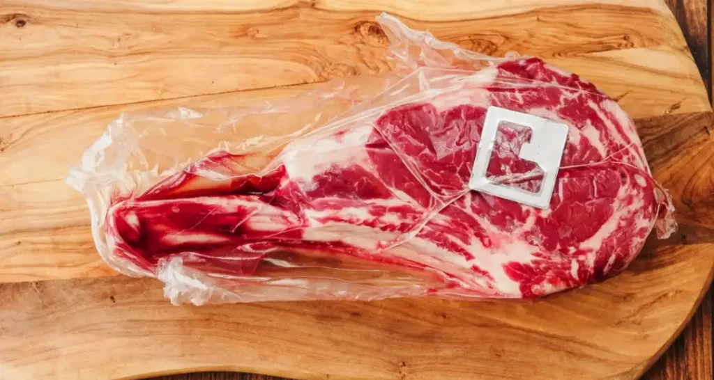 Carne de boi em peça, com gordura e carne, embalado a vácuo sobre tábua de madeira.