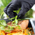 A imagem mostra uma mão usando uma luva preta de cozinha enquanto finaliza um prato com cuidado. A pessoa está colocando tiras de ervas frescas (provavelmente cebolinha ou manjericão) sobre um pedaço de salmão grelhado, que tem aparência suculenta e dourada.