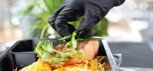 A imagem mostra uma mão usando uma luva preta de cozinha enquanto finaliza um prato com cuidado. A pessoa está colocando tiras de ervas frescas (provavelmente cebolinha ou manjericão) sobre um pedaço de salmão grelhado, que tem aparência suculenta e dourada.