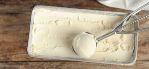 Imagem de um recipiente com sorvete de creme e uma colher de sorvete, em uma superfície de madeira.