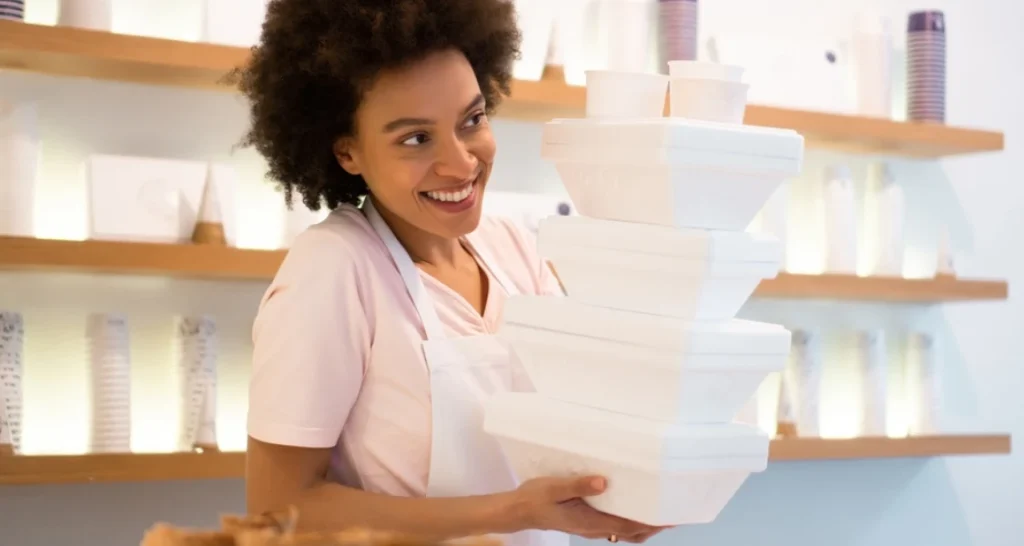 Mulher carregando caixas de alimentos ou produtos em uma loja ou mercado, ambiente bem iluminado.