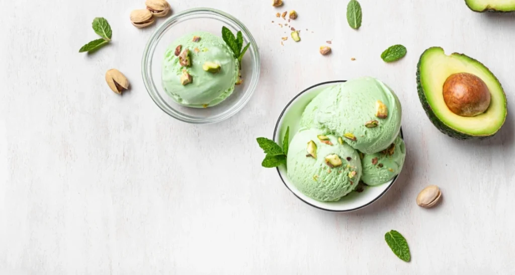 Sorvete de abacate com pistaches e hortelã, servido em taças transparentes, ao lado de abacate fresco, pistaches e folhas de hortelã na mesa branca.