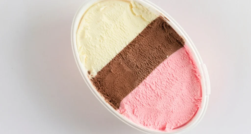 Sorvete de três sabores diferentes: morango, chocolate e creme, em um copo plástico ovalado sobre fundo branco.