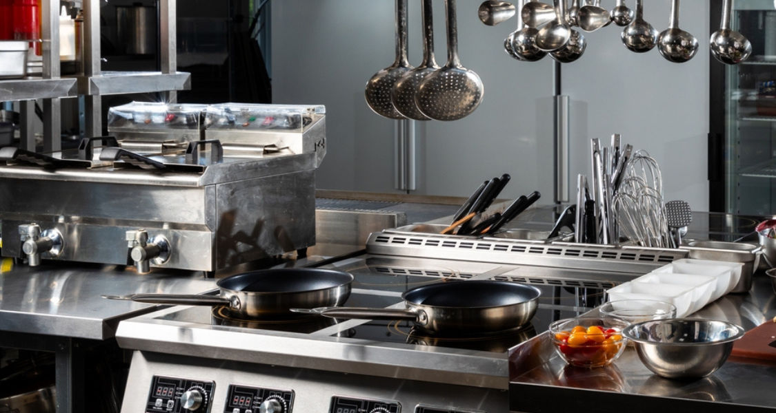 A imagem é uma fotografia em close-up que retrata uma estação de trabalho de cocção e preparo em uma cozinha industrial ou profissional, destacando a organização e os equipamentos de aço inoxidável.