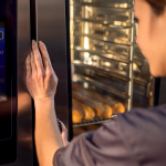 A imagem é uma fotografia em close-up que foca na interação de uma pessoa com um forno combinado industrial moderno ou equipamento similar de alta tecnologia para cozinha profissional.