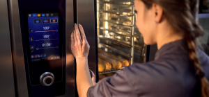 A imagem é uma fotografia em close-up que foca na interação de uma pessoa com um forno combinado industrial moderno ou equipamento similar de alta tecnologia para cozinha profissional.