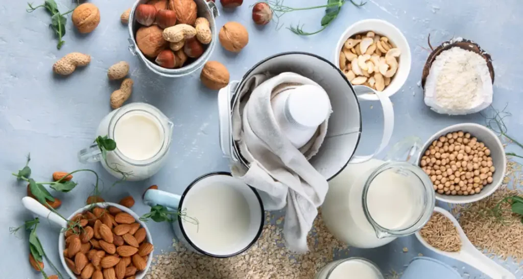 Vários ingredientes de origem vegetal, como castanhas, grão-de-bico, aveia, nozes e leite vegetal, dispostos sobre uma mesa de cor azul claro, ideal para receitas veganas e alimentação natural.
