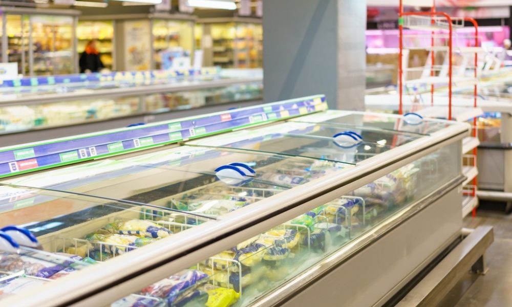Vista em perspectiva de um corredor de supermercado focado em ilhas horizontais e balcões refrigerados de vidro repletos de produtos congelados.