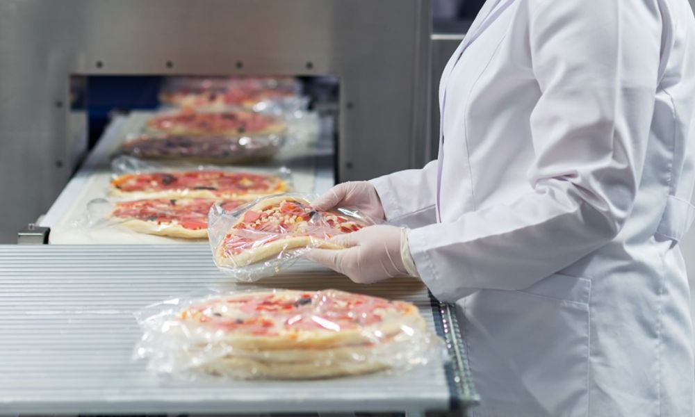 Linha de produção industrial onde um operador de jaleco branco e luvas manuseia pizzas embaladas a vácuo em uma esteira rolante.