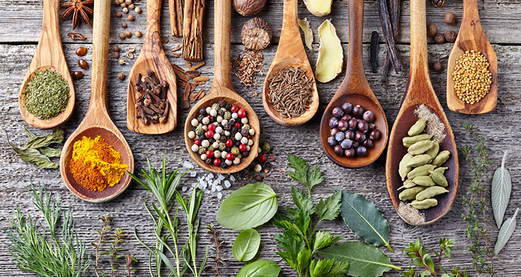 Colheres de madeira posicionadas sobre uma mesa também de madeira, com diversos tipos de temperos para representar as tendências de sabores para 2023.