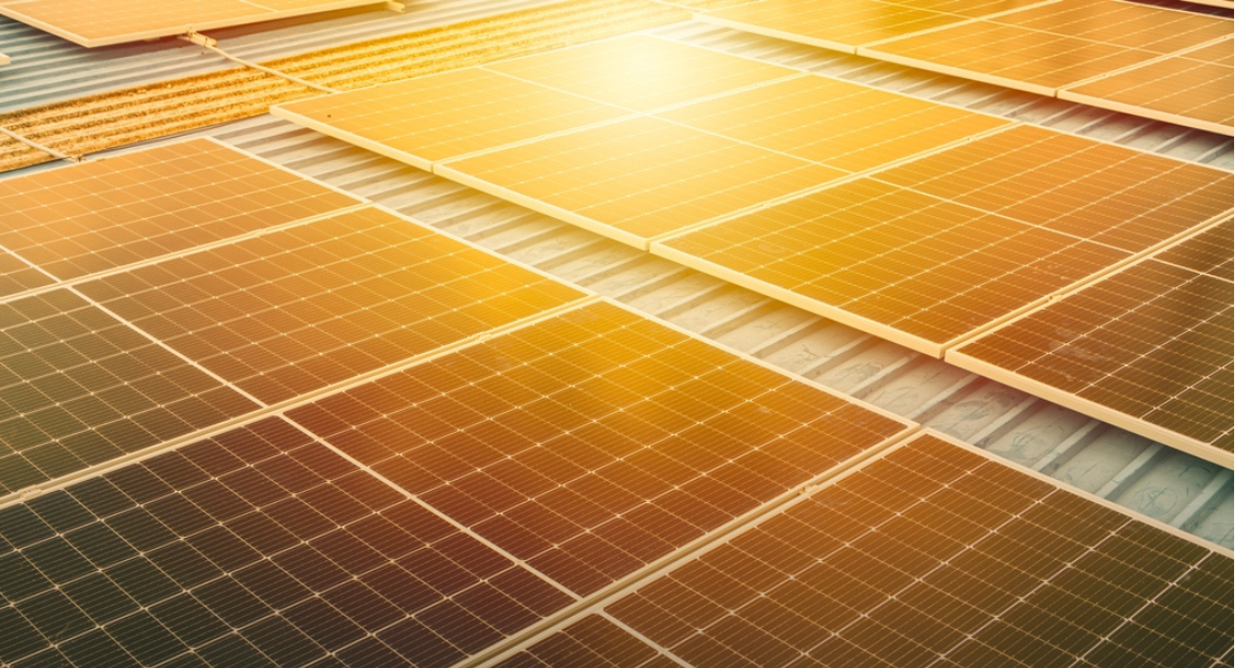 A imagem mostra uma instalação de painéis solares fotovoltaicos em um telhado metálico, provavelmente de um galpão industrial. A iluminação solar intensa, com tons dourados e alaranjados, destaca o brilho e a eficiência da captação de energia solar pelos painéis. O efeito de luz reforça visualmente a intensidade da radiação solar, que é essencial para a produção de energia limpa e renovável.