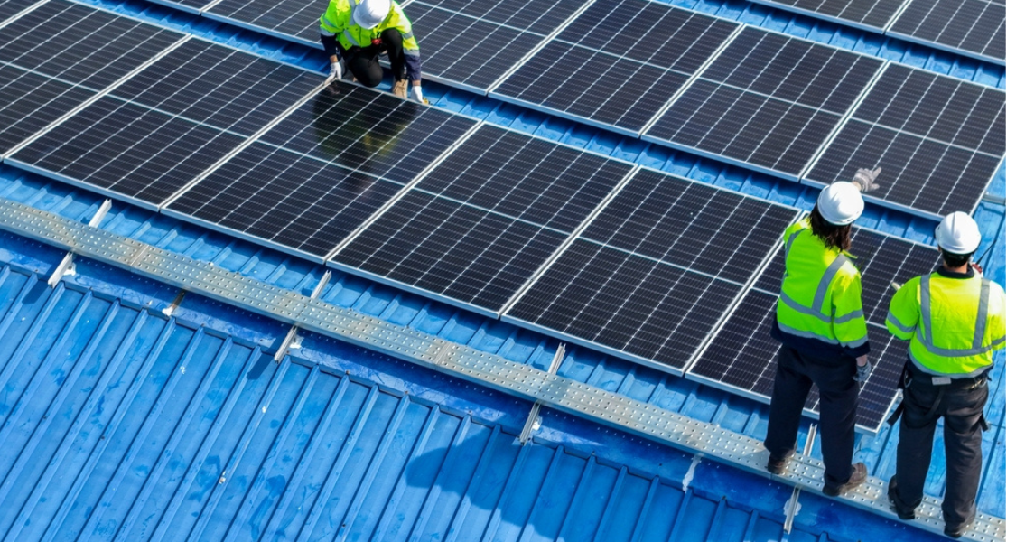 A imagem mostra trabalhadores instalando painéis solares em um telhado metálico azul, provavelmente de um galpão industrial ou comercial. Três pessoas com equipamentos de segurança (capacetes e coletes refletivos) estão envolvidas na atividade: uma está ajoelhada sobre os painéis, provavelmente ajustando ou fixando a estrutura, enquanto duas observam e analisam o progresso, indicando a direção ou discutindo o projeto.