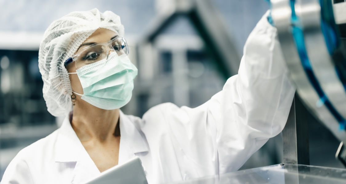 Uma mulher com vestimenta de laboratório (jaleco branco, máscara facial, óculos de segurança e touca descartável) olha atentamente para um equipamento.