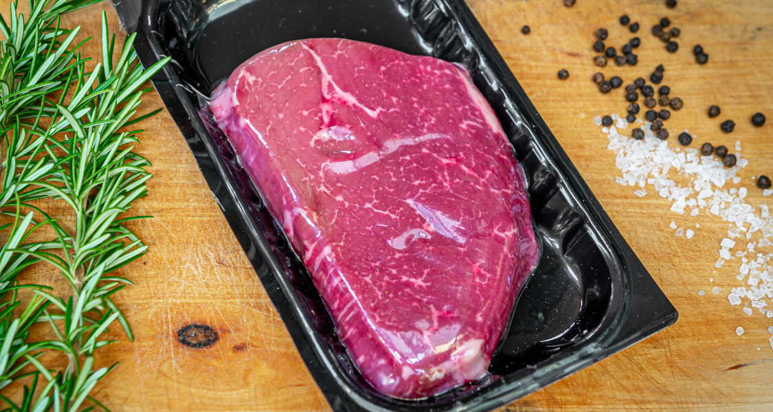 A imagem mostra um pedaço de carne vermelha fresca, embalada a vácuo em uma bandeja plástica preta, colocada sobre uma tábua de madeira.