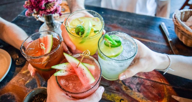 Quatro pessoas brindando com coquetéis coloridos em um ambiente descontraído. As bebidas incluem frutas como melancia e limão, ideal para festas e encontros.