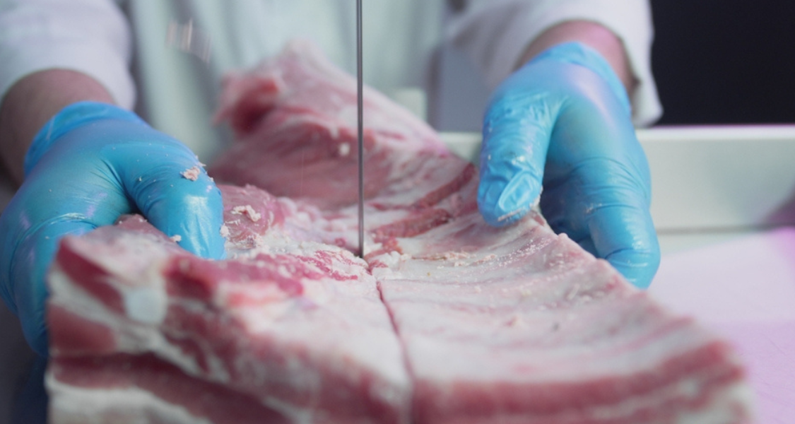 A imagem é um close-up (plano detalhe) de uma operação de corte industrial de carne, focando na precisão e nas medidas de higiene.