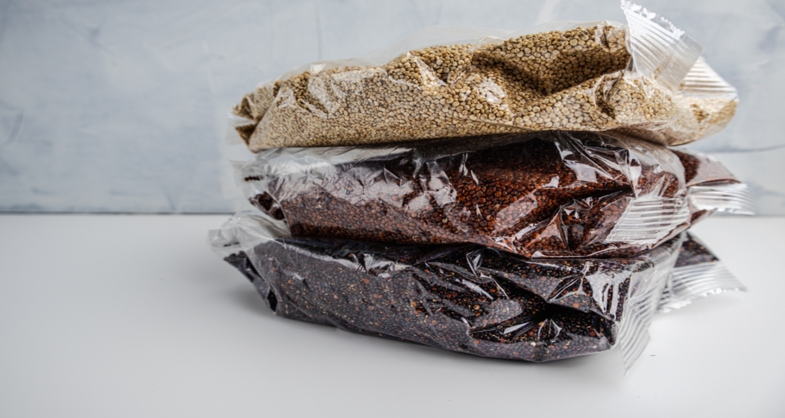 A imagem mostra três pacotes transparentes empilhados, contendo grãos embalados a vácuo ou selados em plástico. No topo, há um pacote com grãos claros e pequenos, semelhantes à quinoa branca. No meio, um pacote com grãos avermelhados, e na base, um com grãos escuros, possivelmente quinoa preta ou outro tipo de semente.