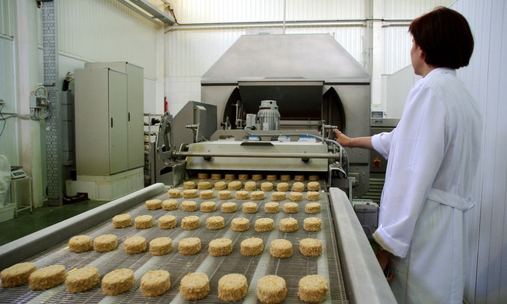Uma mulher vestida com jaleco branco supervisionando a produção industrial de alimentos em uma esteira rolante carregada de hambúrgueres empanados.