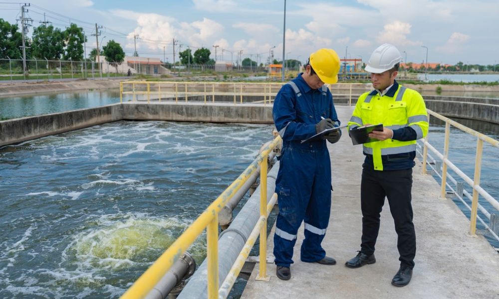 Dois trabalhadores usando capacetes e roupas de proteção inspecionando e anotando dados em uma prancheta ao lado de um tanque de tratamento de água/efluents industrial.