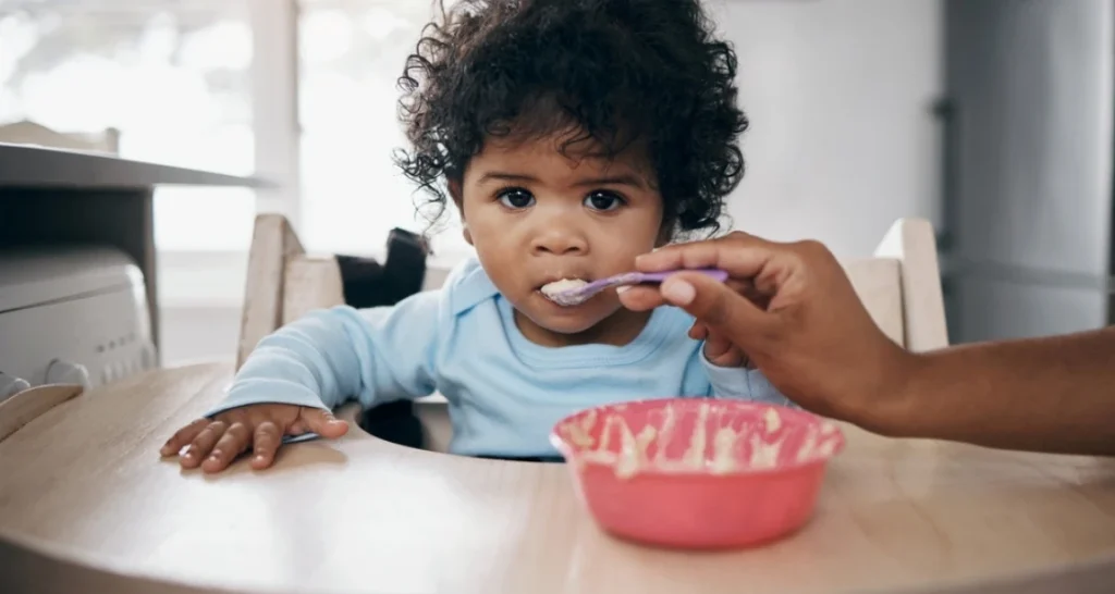 Criança de cabelos cacheados e pele morena criança sendo alimentada com colher, sentado em cadeira de alta sobre uma mesa, em ambiente doméstico.