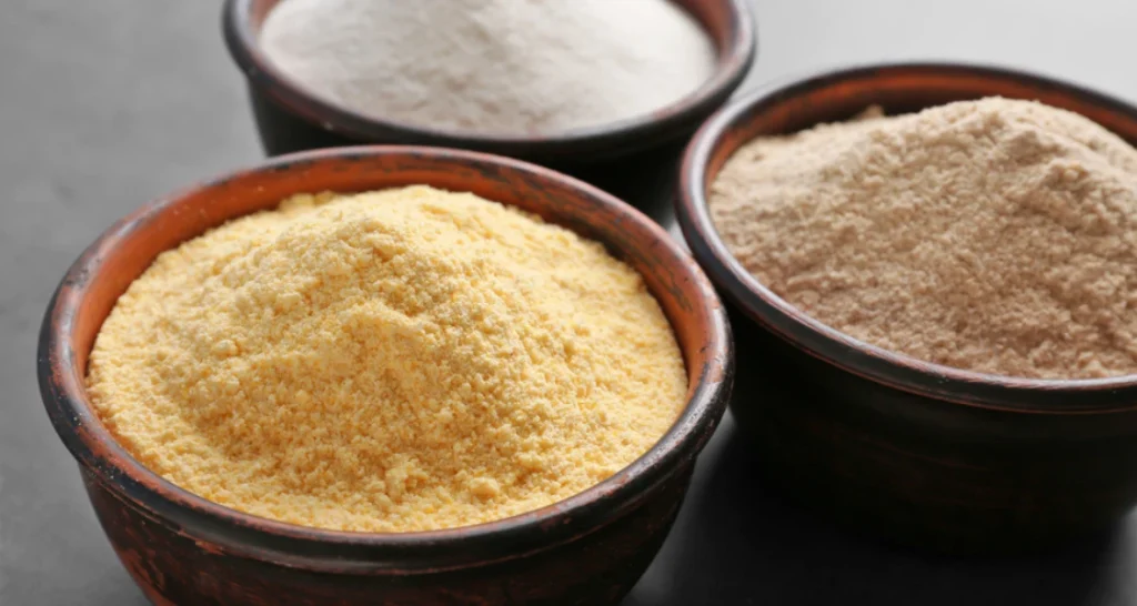 Três tigelas com diferentes tipos de farinha, incluindo farinha de milho, farinha de trigo e farinha de aveia, usadas na culinária brasileira.