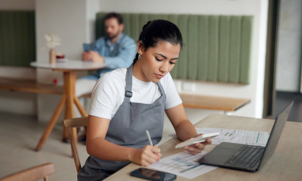 ma funcionária (possivelmente uma gerente ou proprietária) concentrada, usando uma calculadora, notebook e papéis. Isso ilustra o processo administrativo de calcular as retenções e a distribuição das gorjetas