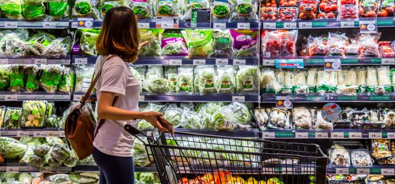 Uma mulher observa as prateleiras de vegetais e hortaliças em um supermercado, com um carrinho de compras ao seu lado. Esta imagem destaca a variedade de produtos frescos disponíveis.