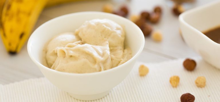 Delicioso sorvete de banana servido em uma tigela branca, acompanhado por frutas secas e um molho escuro, ideal para dias quentes e sobremesas saudáveis.